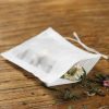 Sachet infuseur en tissu non tissé contenant des plantes sèches, posé sur une table en bois avec des fragments de plantes autour.