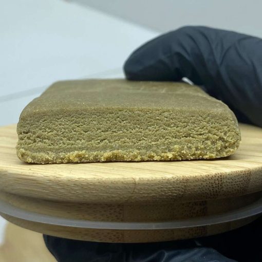 Bloc de résine de cannabis 3x filtré, texture fine et homogène, posé sur un plateau en bois, tenu par une main gantée.